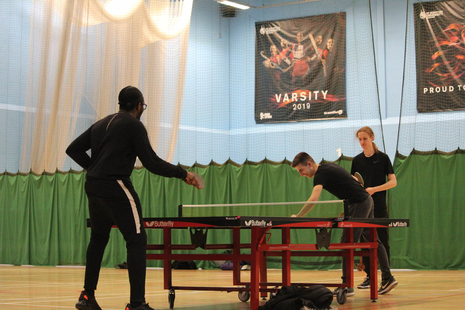 Three people playing table tennis indoors with green barriers and posters on the wall in the background.