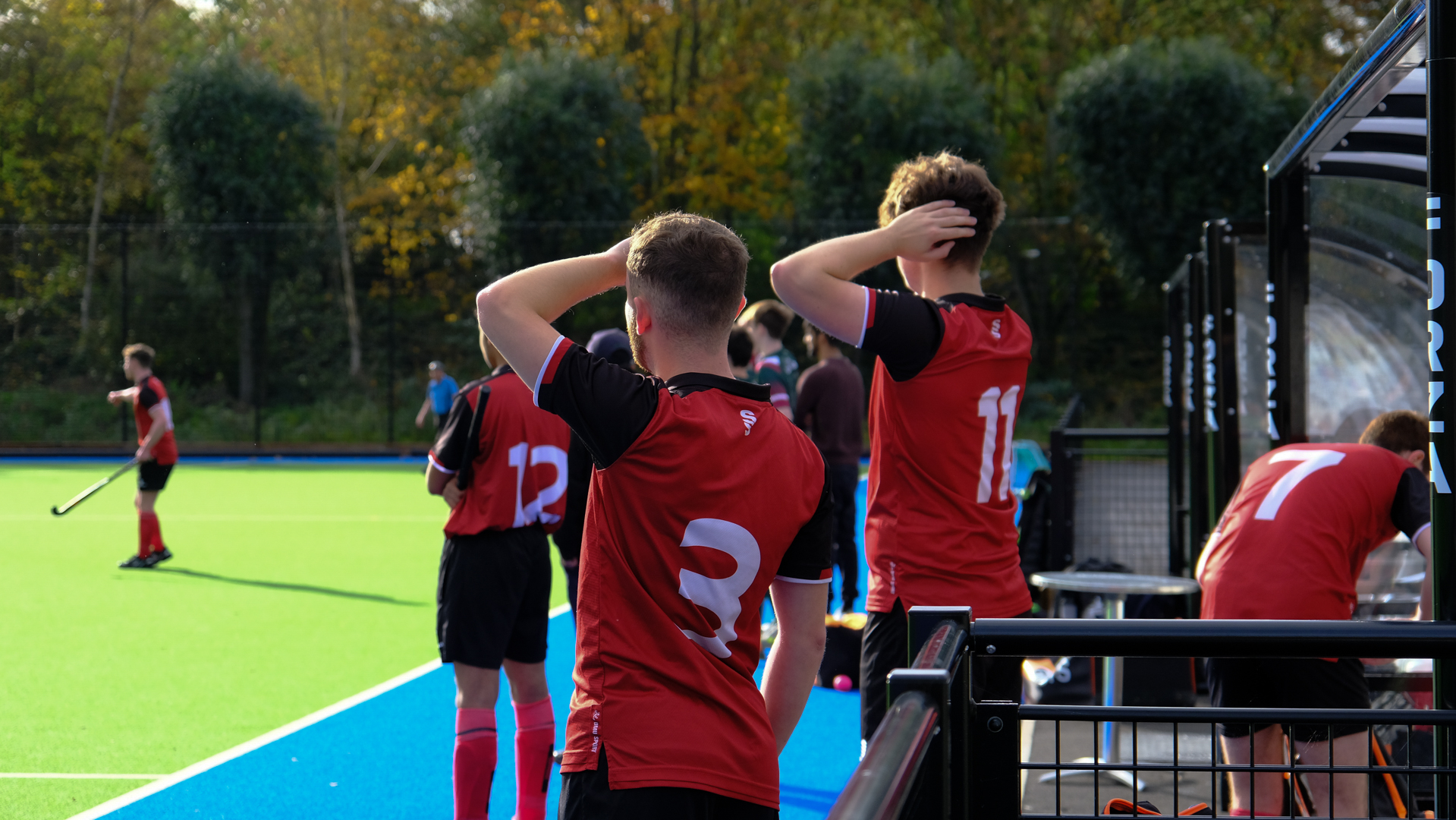 Hockey players in red and black kits stand on the sideline, watching the game.