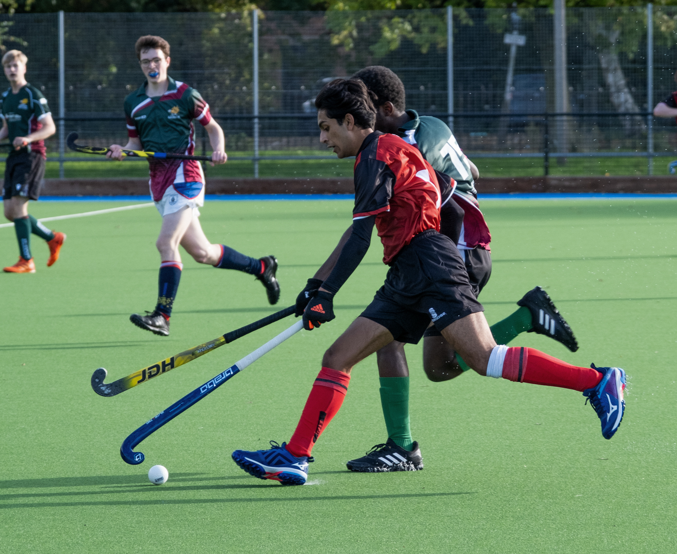 Hockey players in action, with one player dribbling the ball while others move nearby on a green pitch.