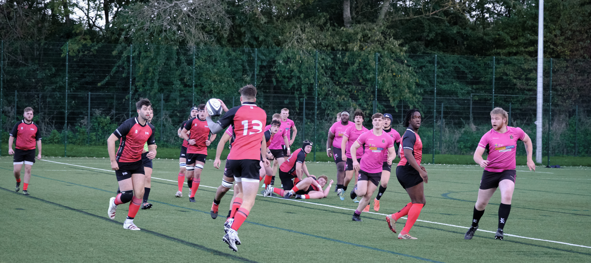 Rugby players in red and pink jerseys competing on a grassy field, with some players running and others on the ground.