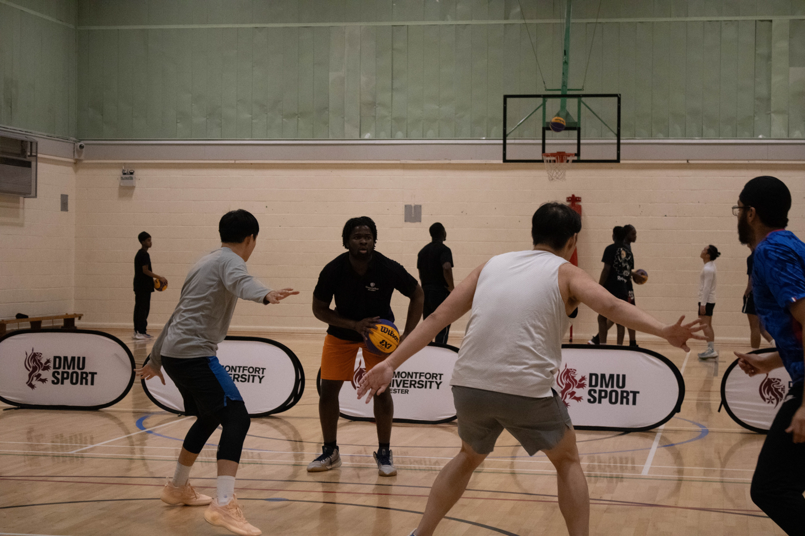 Four people playing basketball indoors, with a hoop and other players in the background.