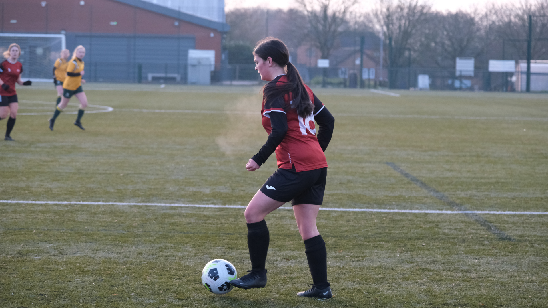 Football player in a red and black jersey controls the ball on a grassy field during a match.