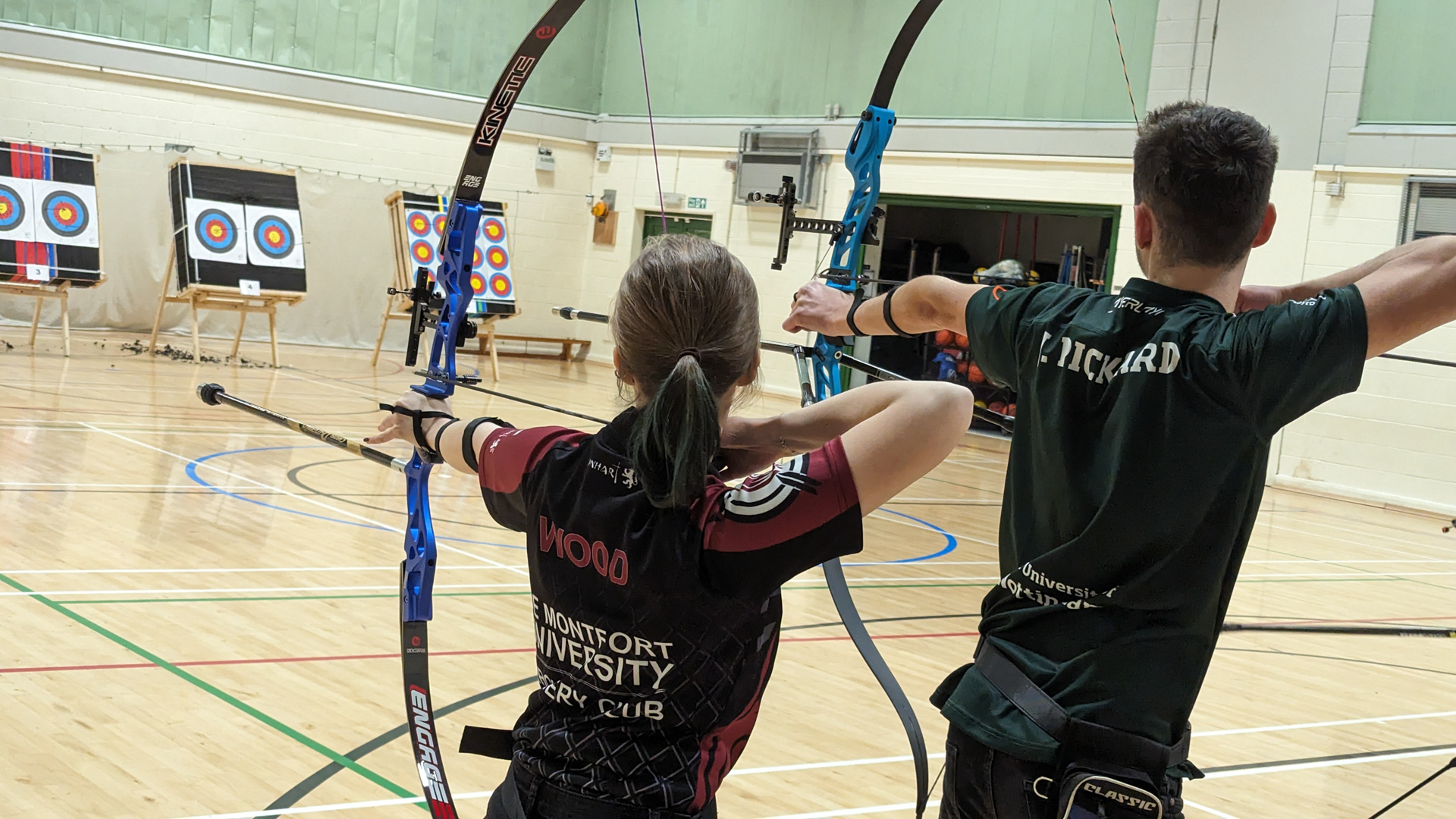 Two archers draw their bows in a gymnasium, aiming at targets set up across the room.