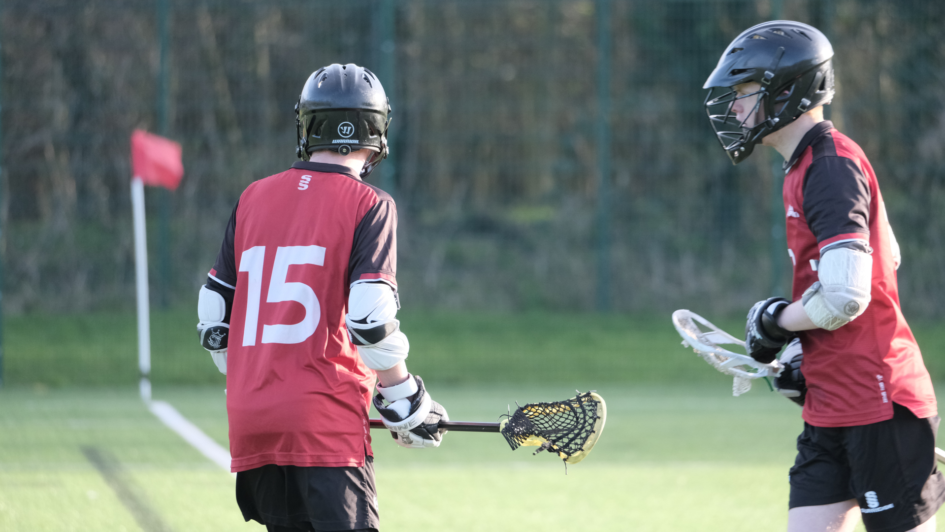 Two lacrosse players in red jerseys and helmets on a grassy field, one holding lacrosse sticks.