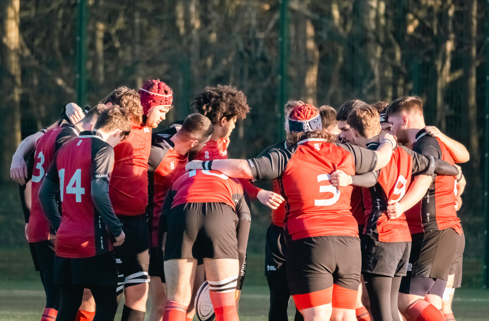 A rugby team in red and black uniforms stands in a huddle on a grassy field.