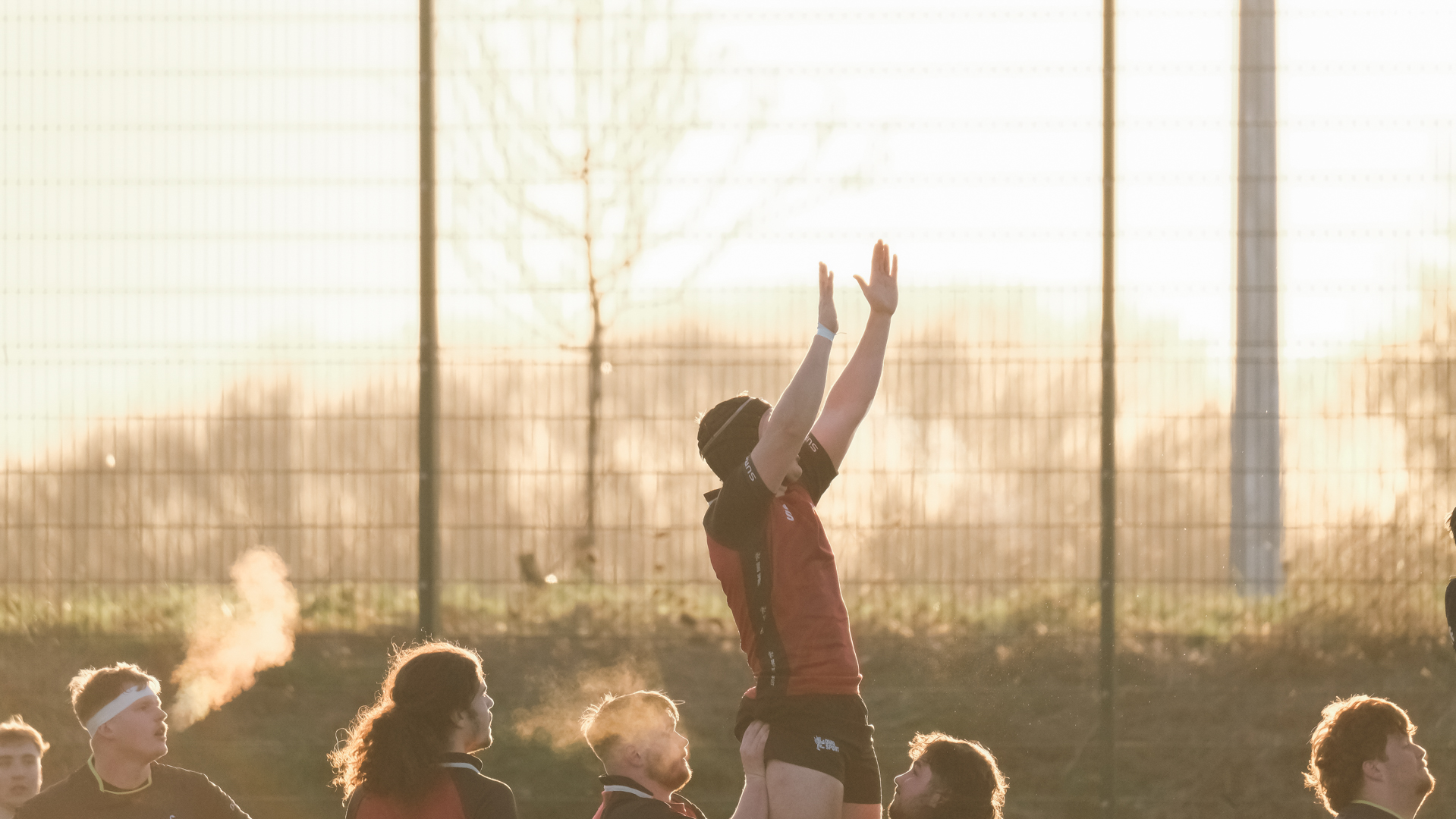 Rugby players on a field, with one player jumping to catch the ball during a match at sunset.