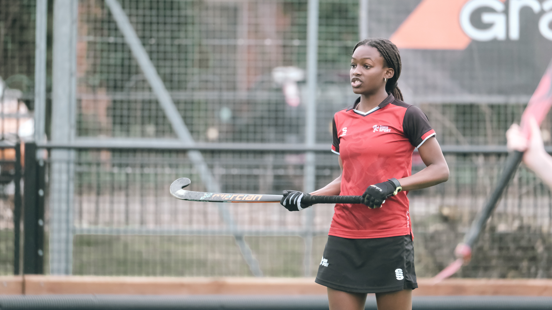 Hockey player in red and black kit holding a hockey stick on an outdoor field.