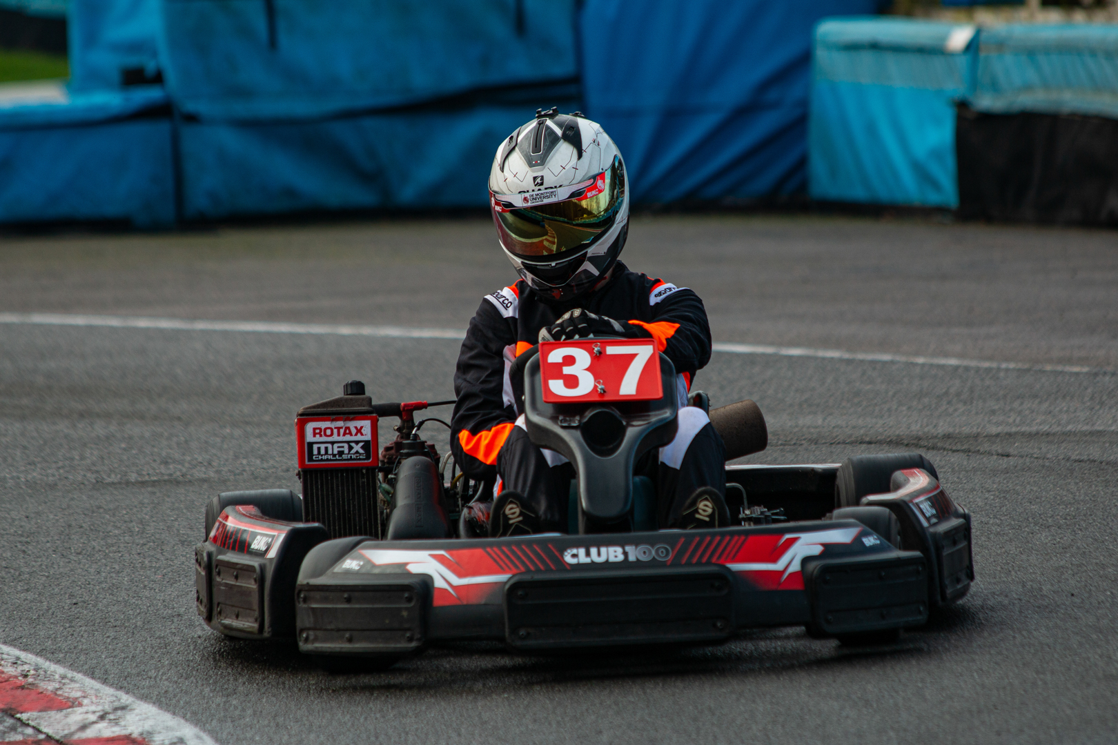 Person wearing a helmet and racing suit driving a go-kart with the number 37 on a track.