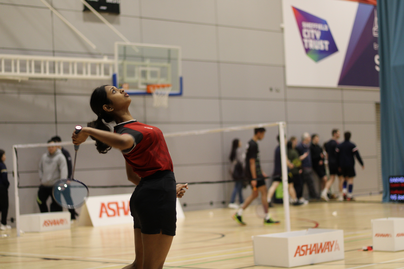 A badminton player in mid-swing on an indoor court, with spectators and other players in the background.