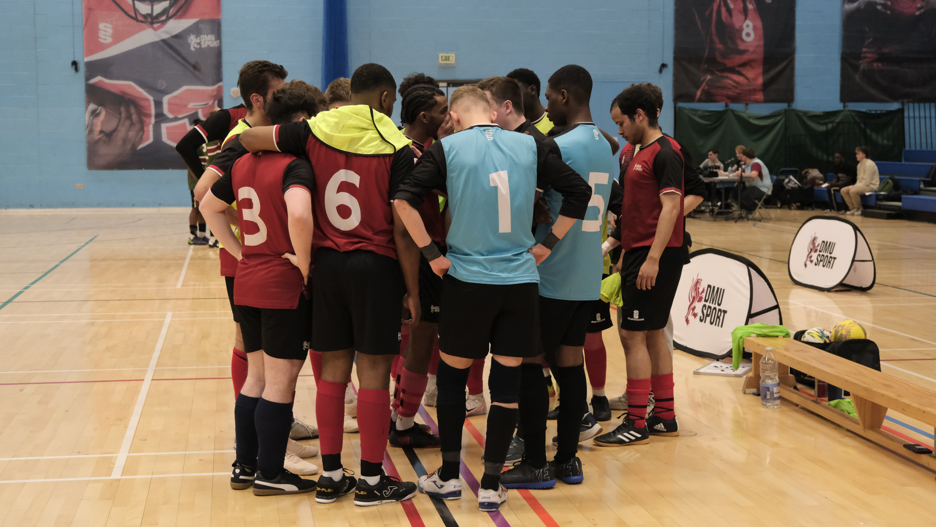 Futsal team huddles together on a gym floor, wearing matching shirts and listening to a coach.