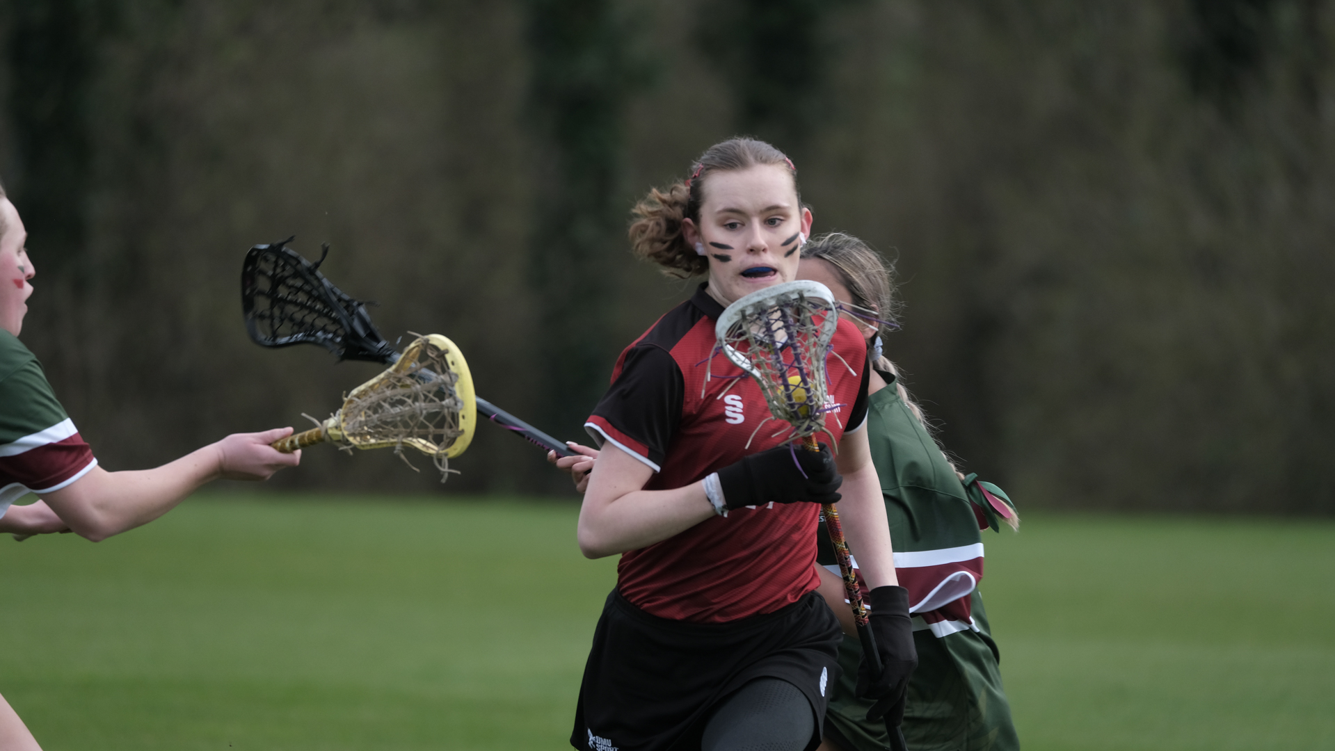 Player in protective gear running with a lacrosse stick during a game on a grassy field.