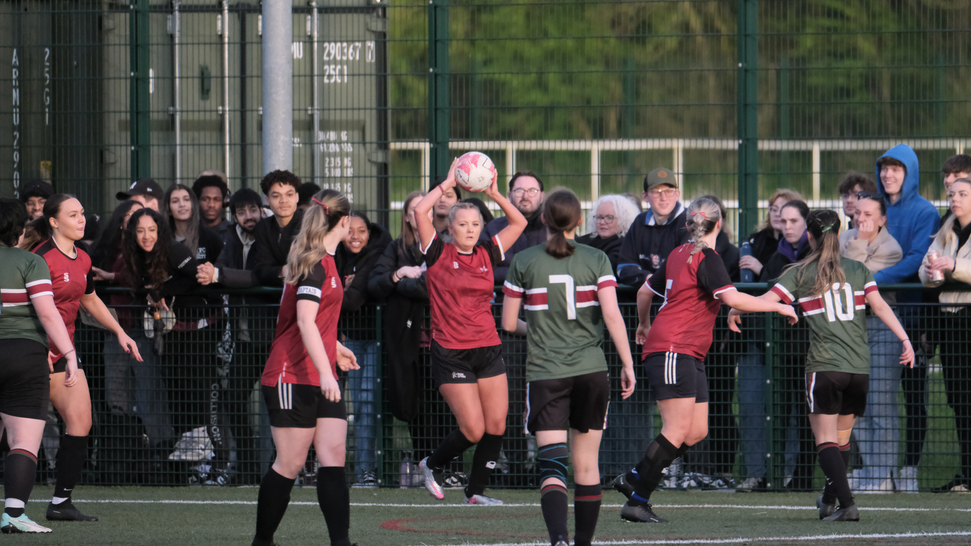 Women’s football match in progress, with player taking a throw-in and spectators watching from the sidelines.