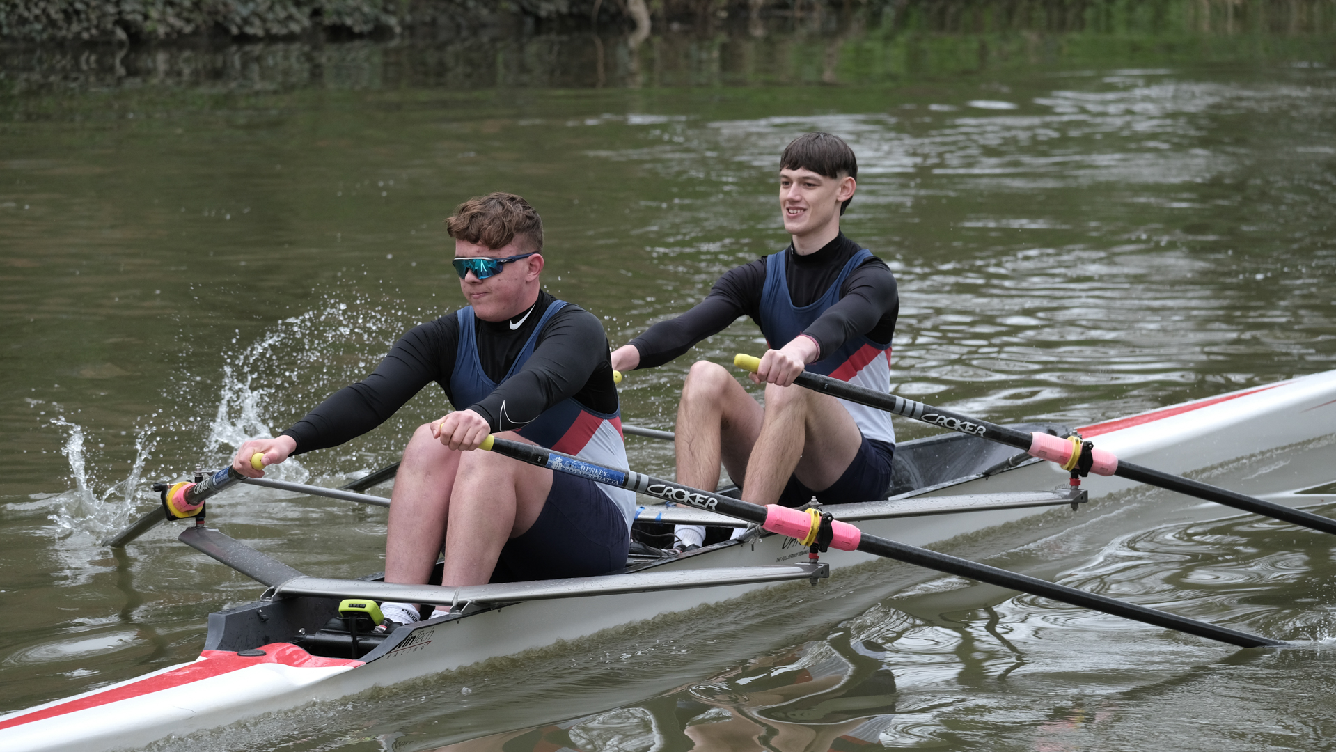 Two people rowing together in a narrow boat on a calm river, wearing matching athletic gear.
