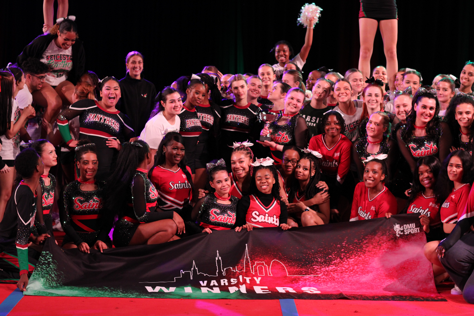 Cheerleading teams posing together on a mat, holding a large banner and celebrating at a competition event.
