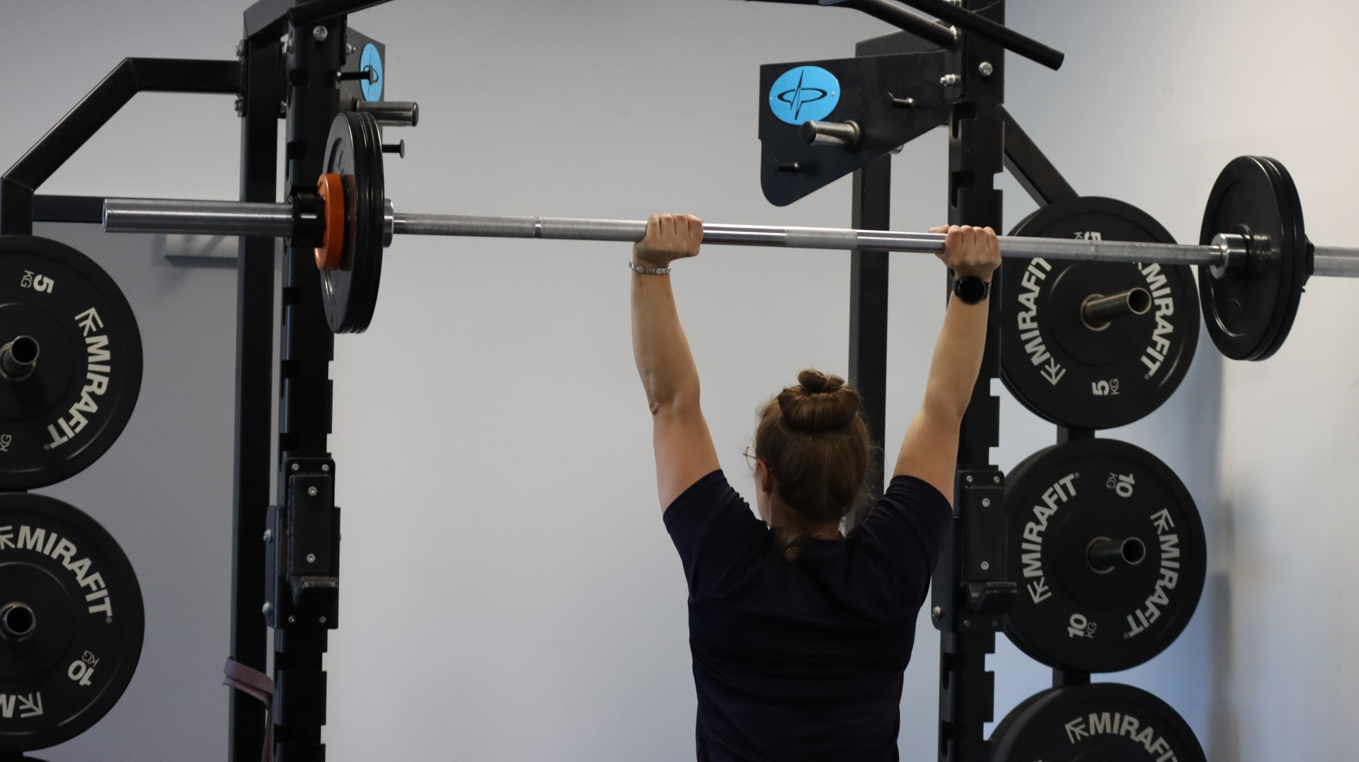 Person lifting a barbell overhead in a gym with weight plates visible on racks nearby.