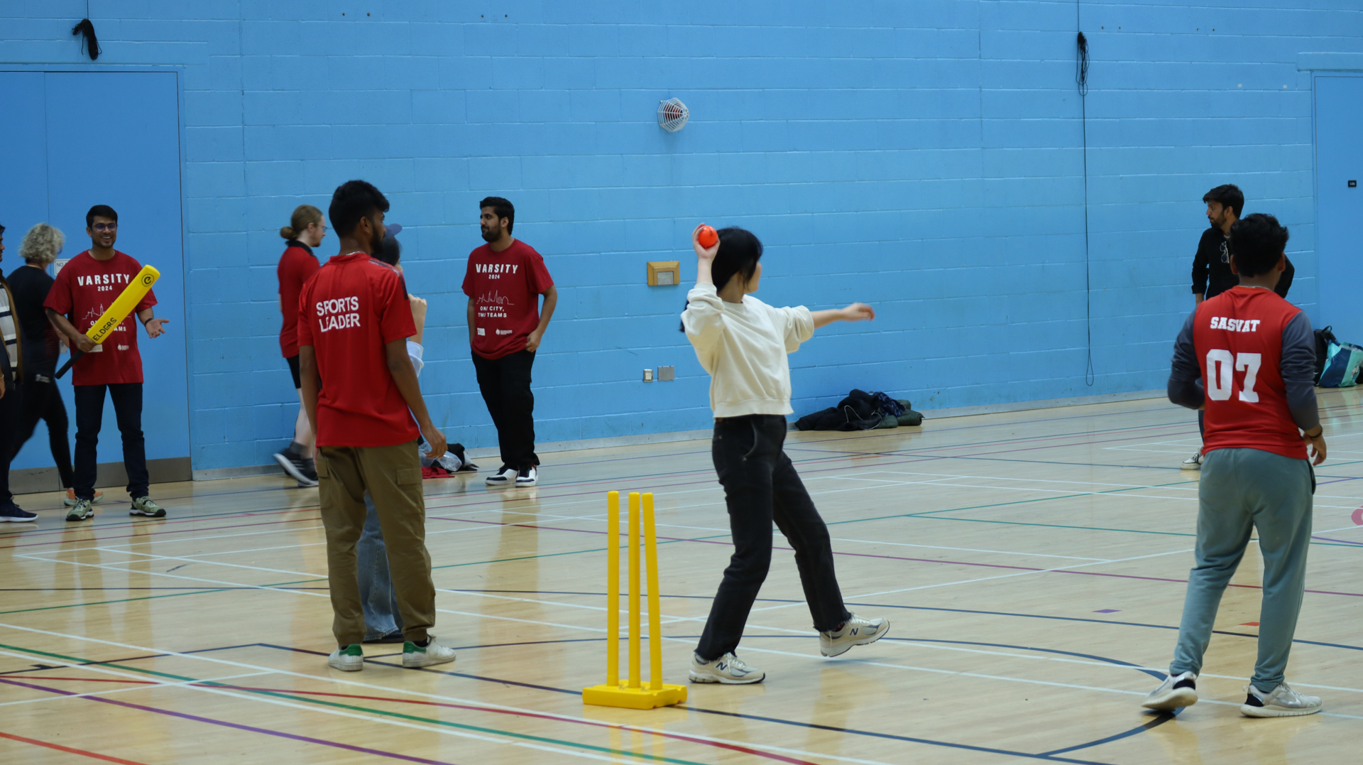Students playing an indoor cricket game, with one student bowling and others fielding or watching.