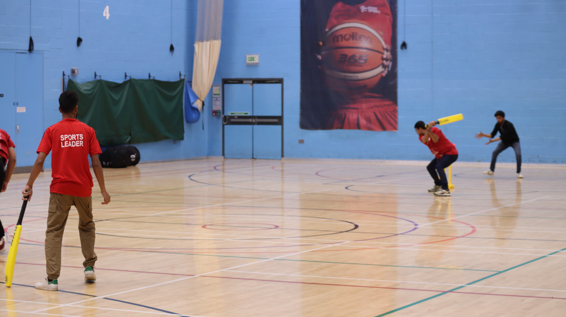 People playing an indoor cricket game with bats and a ball on a gymnasium court.