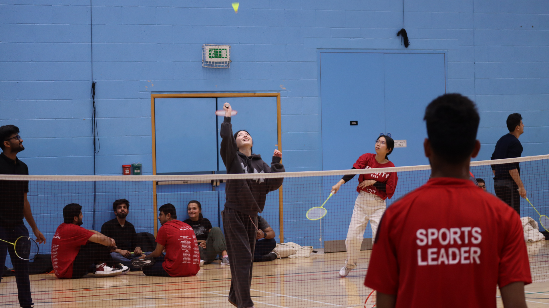 Players participate in badminton in an indoor gym, with others watching.