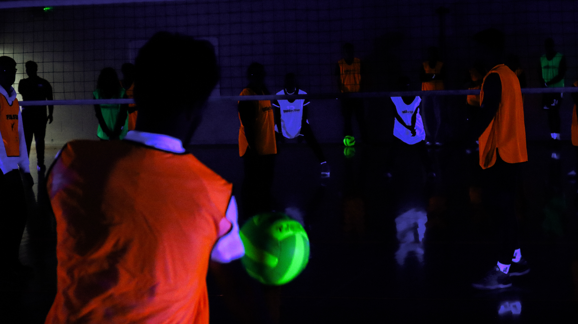 People wearing neon clothing and accessories playing volleyball under neon light in a dark room.
