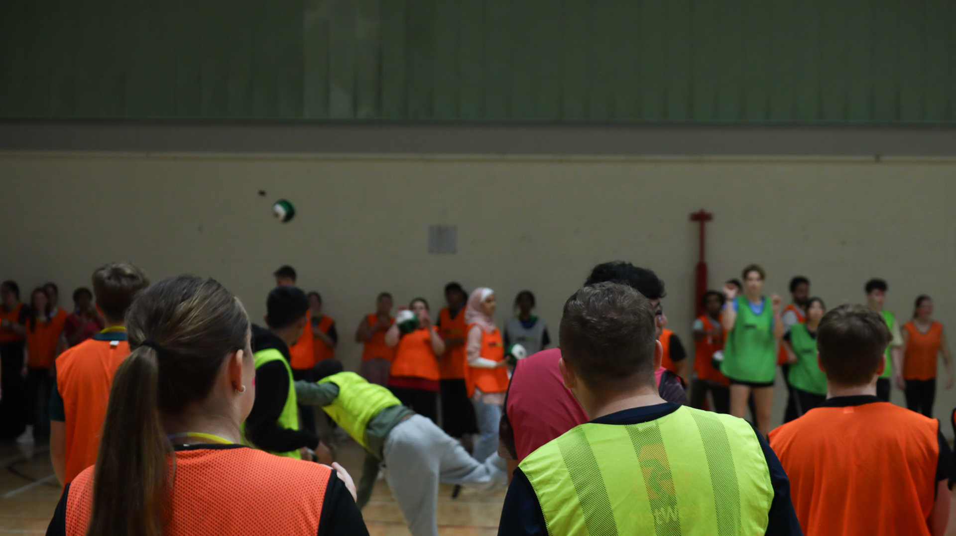A group of people wearing colored vests participate in an indoor dodgeball activity.