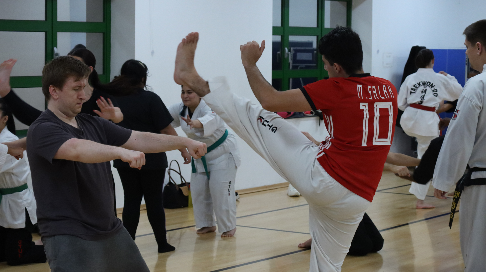 People practicing martial arts in a gym, with one person kicking while others observe or perform techniques.