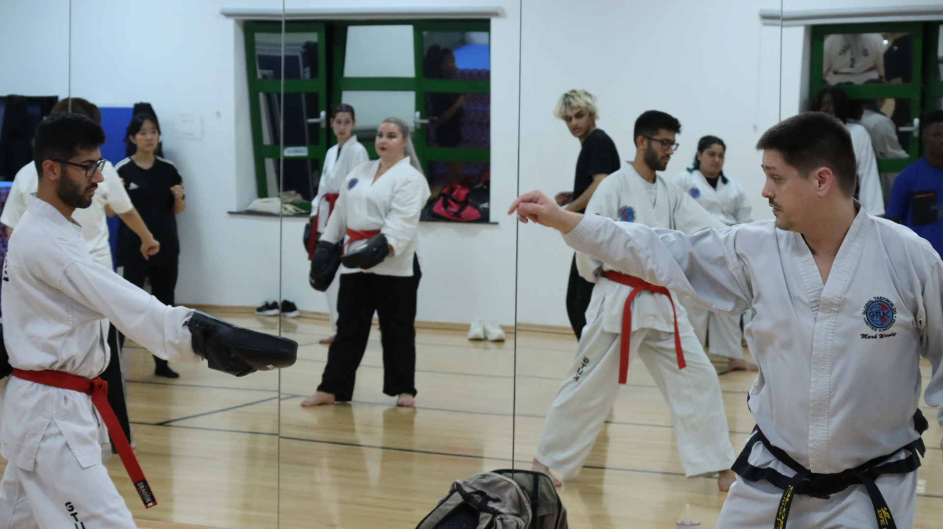 Martial arts students practicing in a gym, with two people sparring in the foreground and others observing.