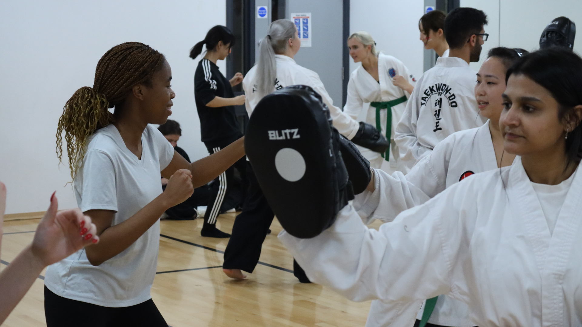 Women practicing martial arts in a gym, with one holding a pad while another prepares to hit it.