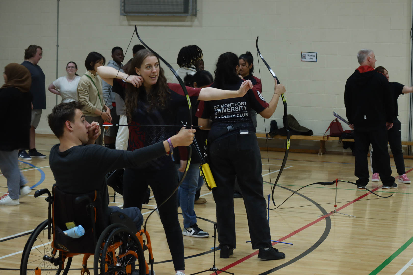 People practicing archery indoors, including a person in a wheelchair aiming a bow.