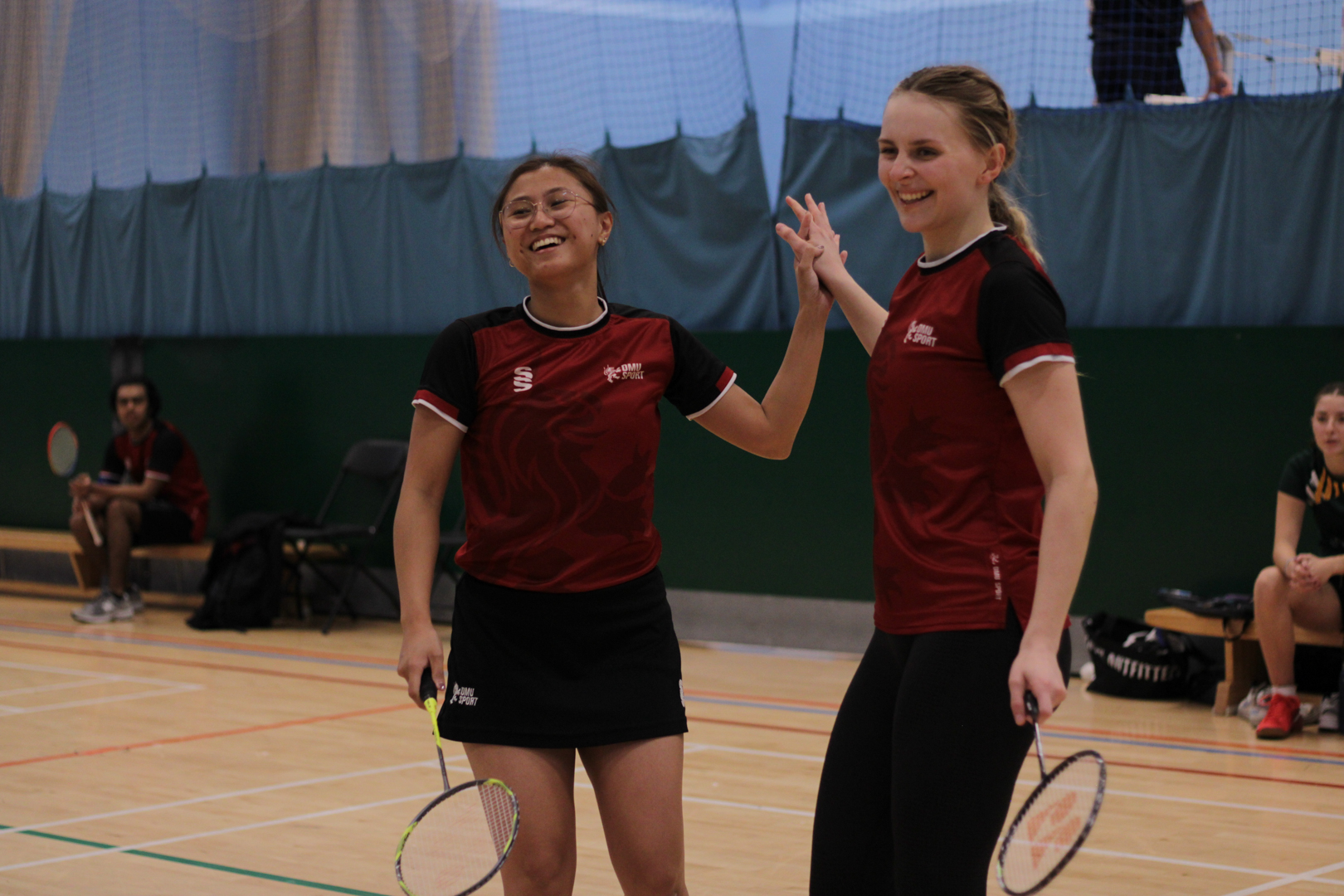 Two female badminton players in matching kits smiling and giving a high five on an indoor court.