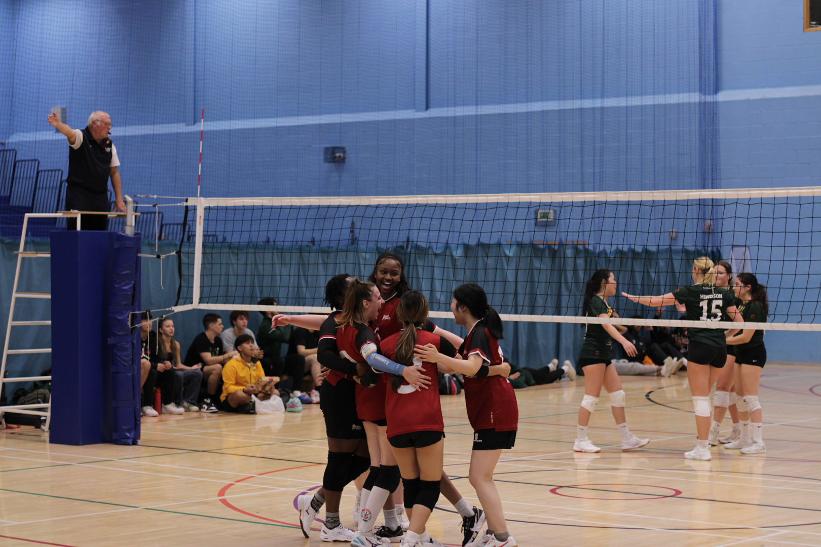 Volleyball teams on an indoor court, with one team celebrating a point while the referee observes from a stand.