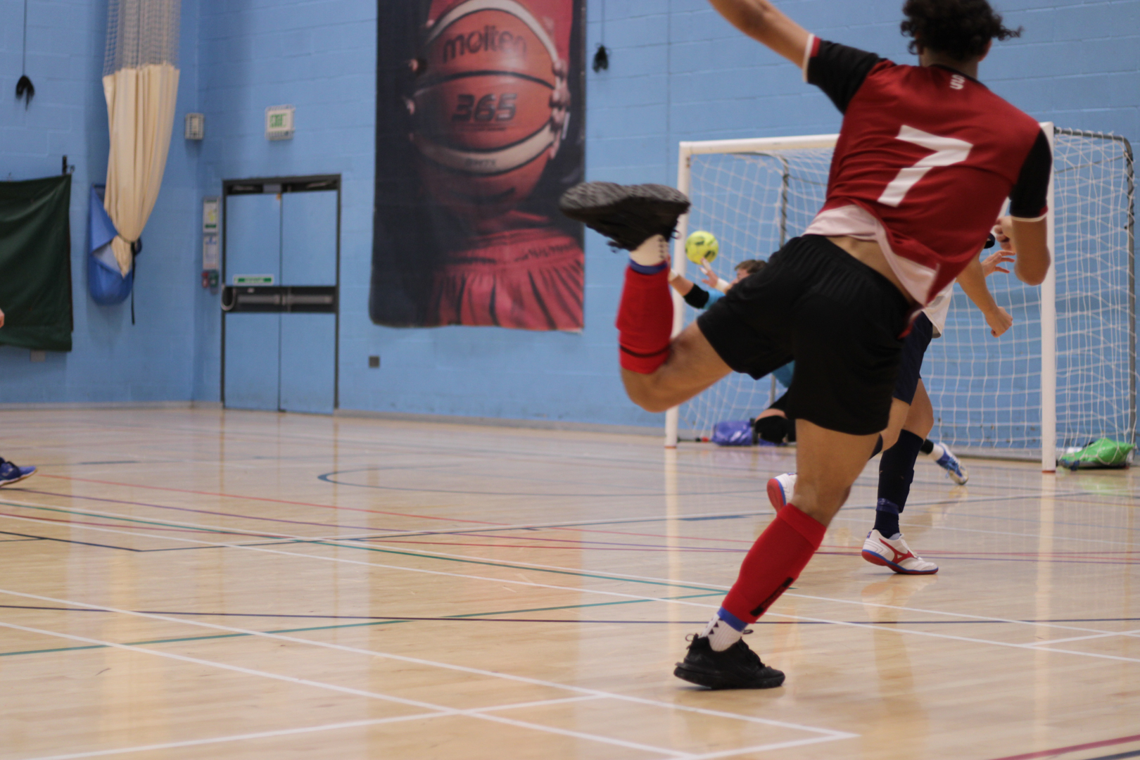 Futsal player in a red jersey kicking a ball into net on an indoor court.
