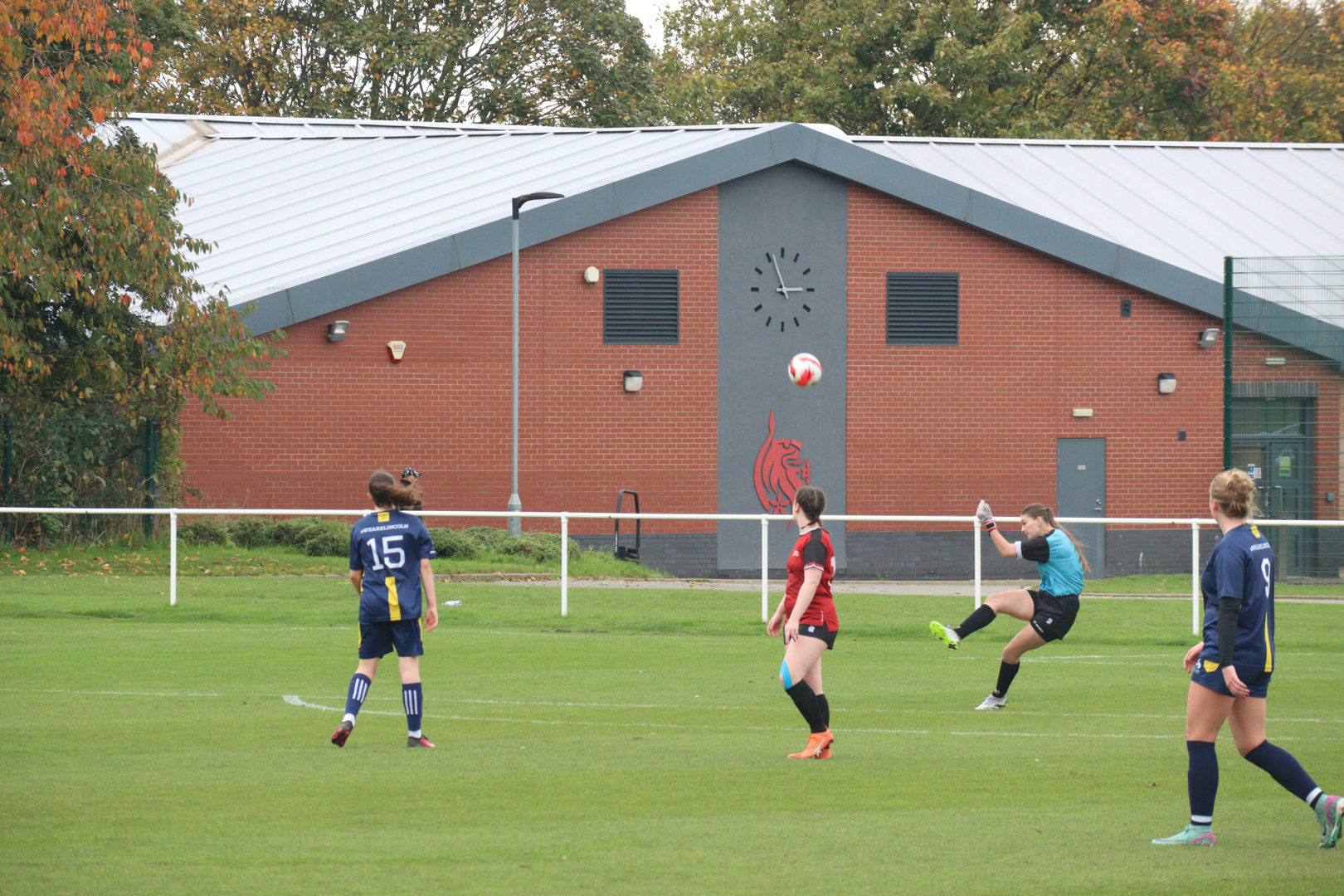 Goalkeeper taking a goal kick during a game, with a brick building in the background.