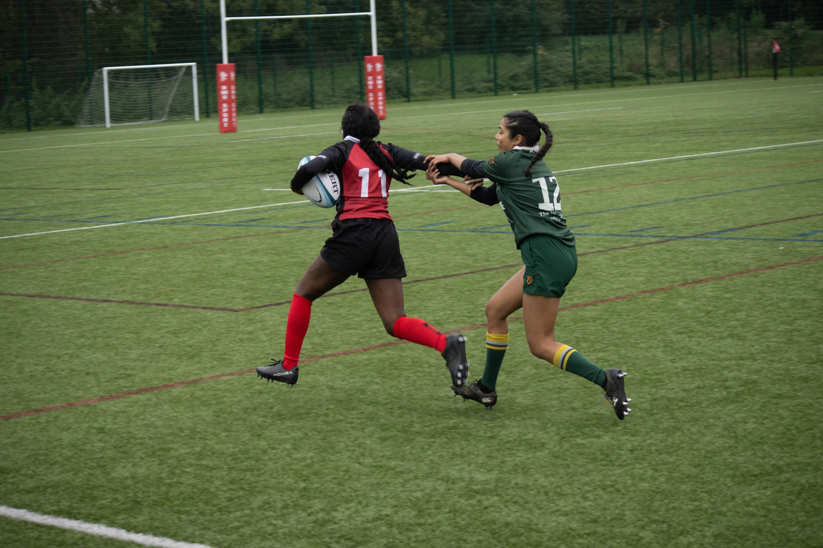 Two Rugby players compete for the ball on a grassy field, one in red and black, the other in green.