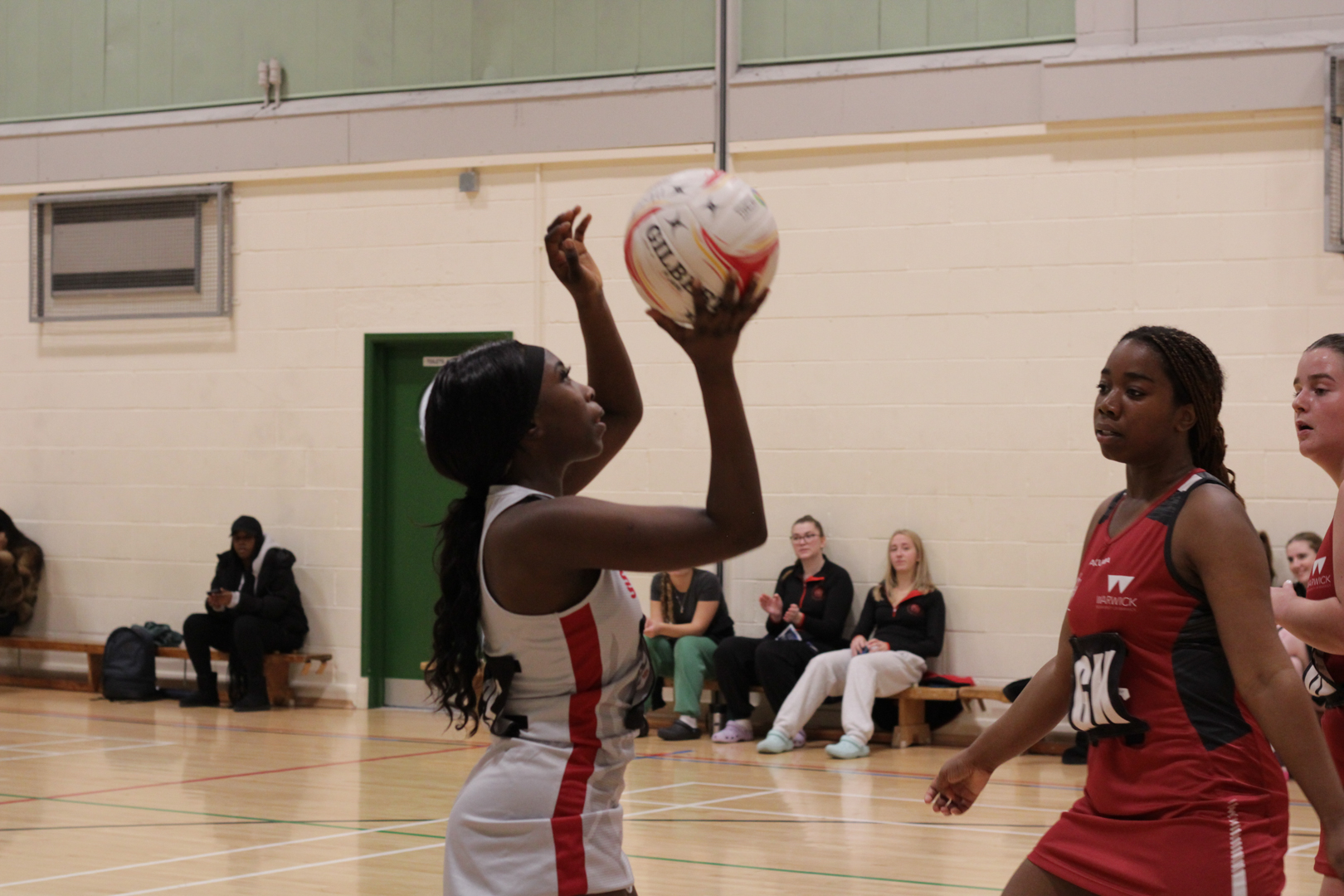 Basketball player in grey preparing to shoot while being guarded by a player in red during a game in a gymnasium.