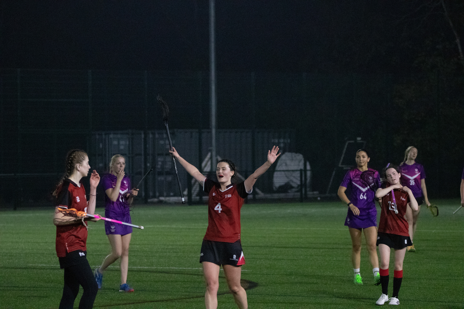 Women playing Lacrosse, with one player in a red and black uniform raising her arms in celebration.