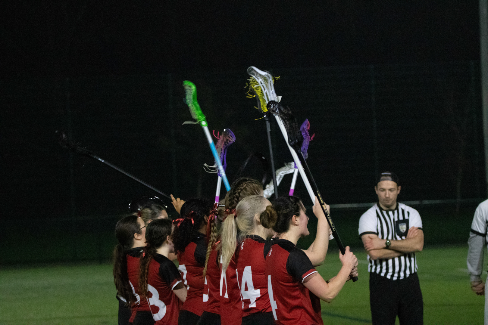 Girls’ lacrosse team in red jerseys raising sticks together on a field at night.