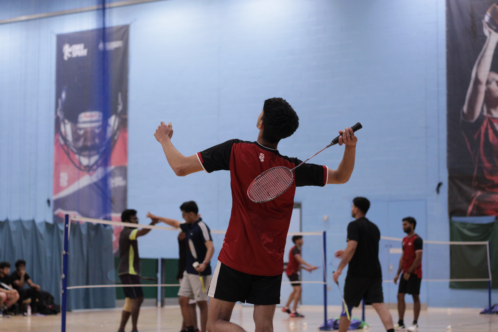 Badminton players in action on an indoor court, with one player preparing to hit the shuttlecock.