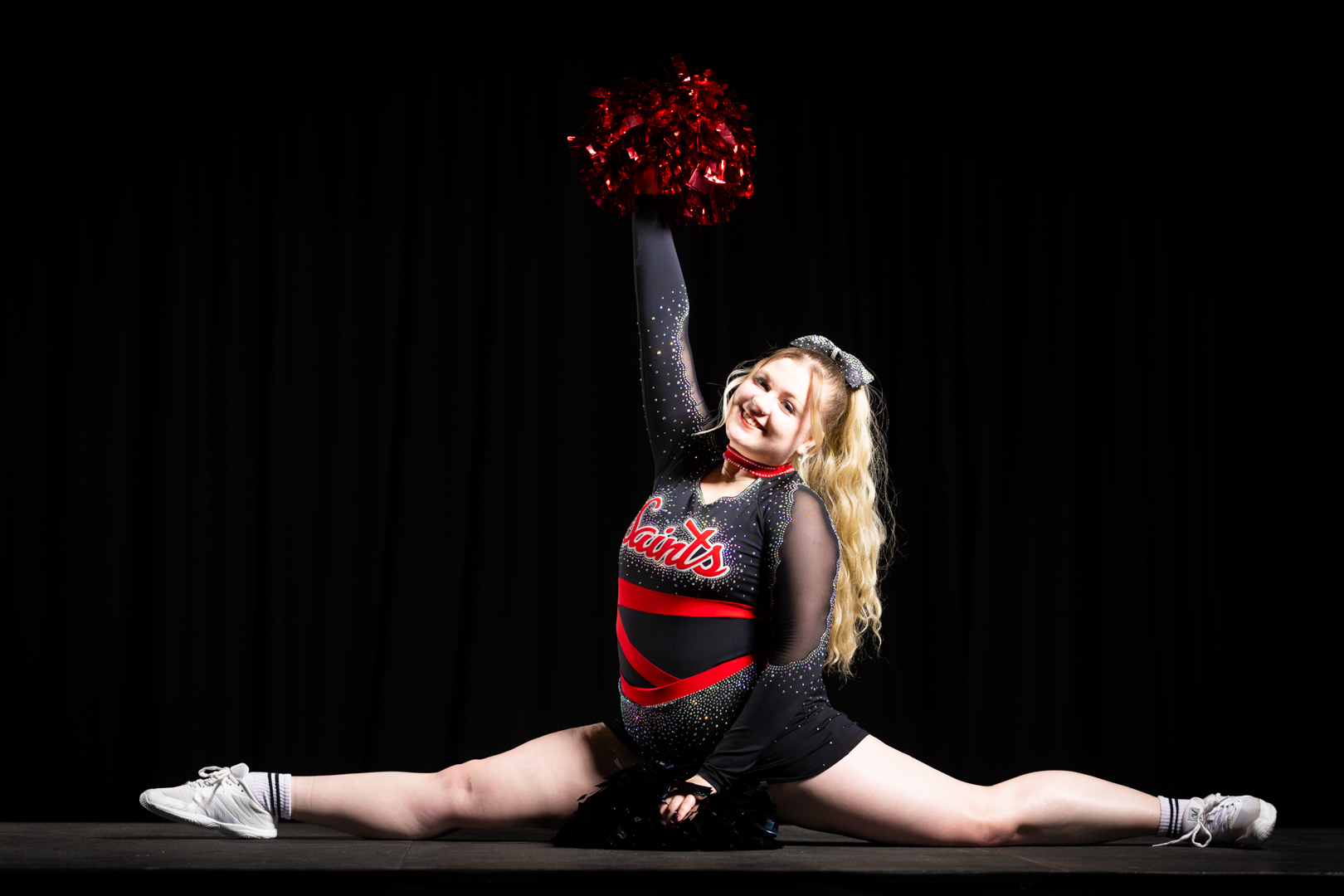 Cheerleader performing a split on stage, holding a red pom-pom up with one arm and smiling.