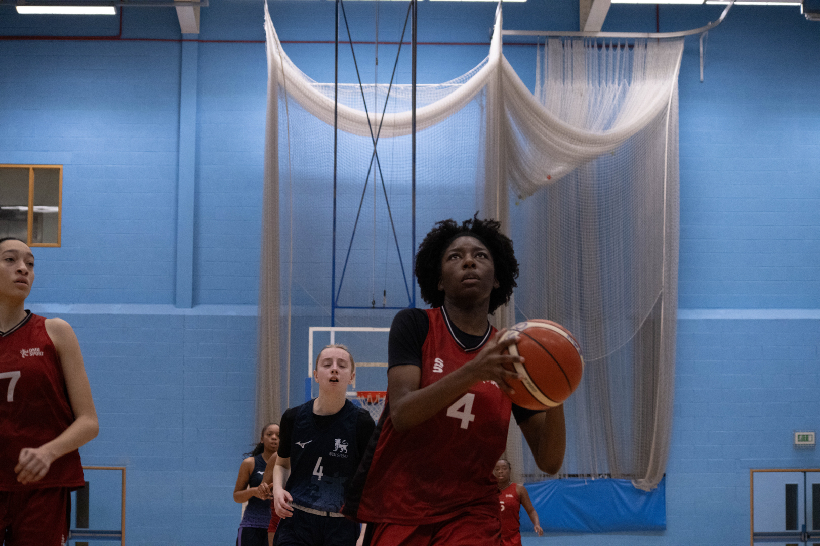 Basketball player in a red kit preparing to shoot while others look on in an indoor gym.
