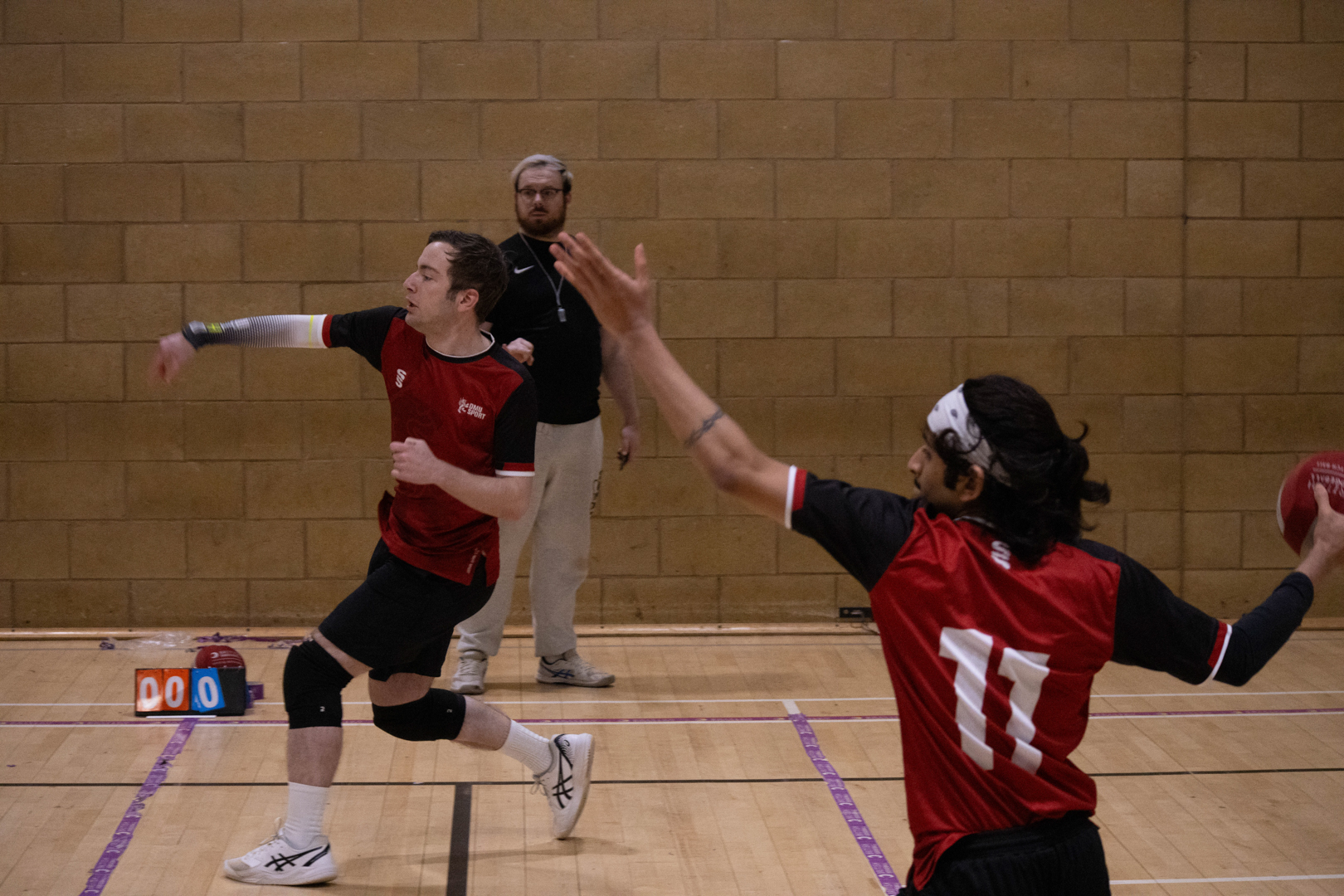 Two athletes play dodgeball on an indoor court, with a referee observing in the background.