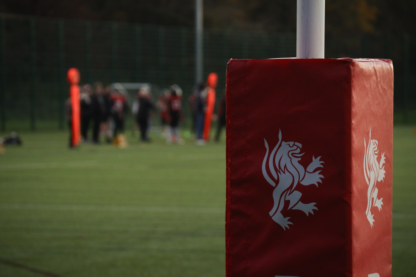 Red goalpost pad with a white lion emblem in focus, with players and field blurred in the background.