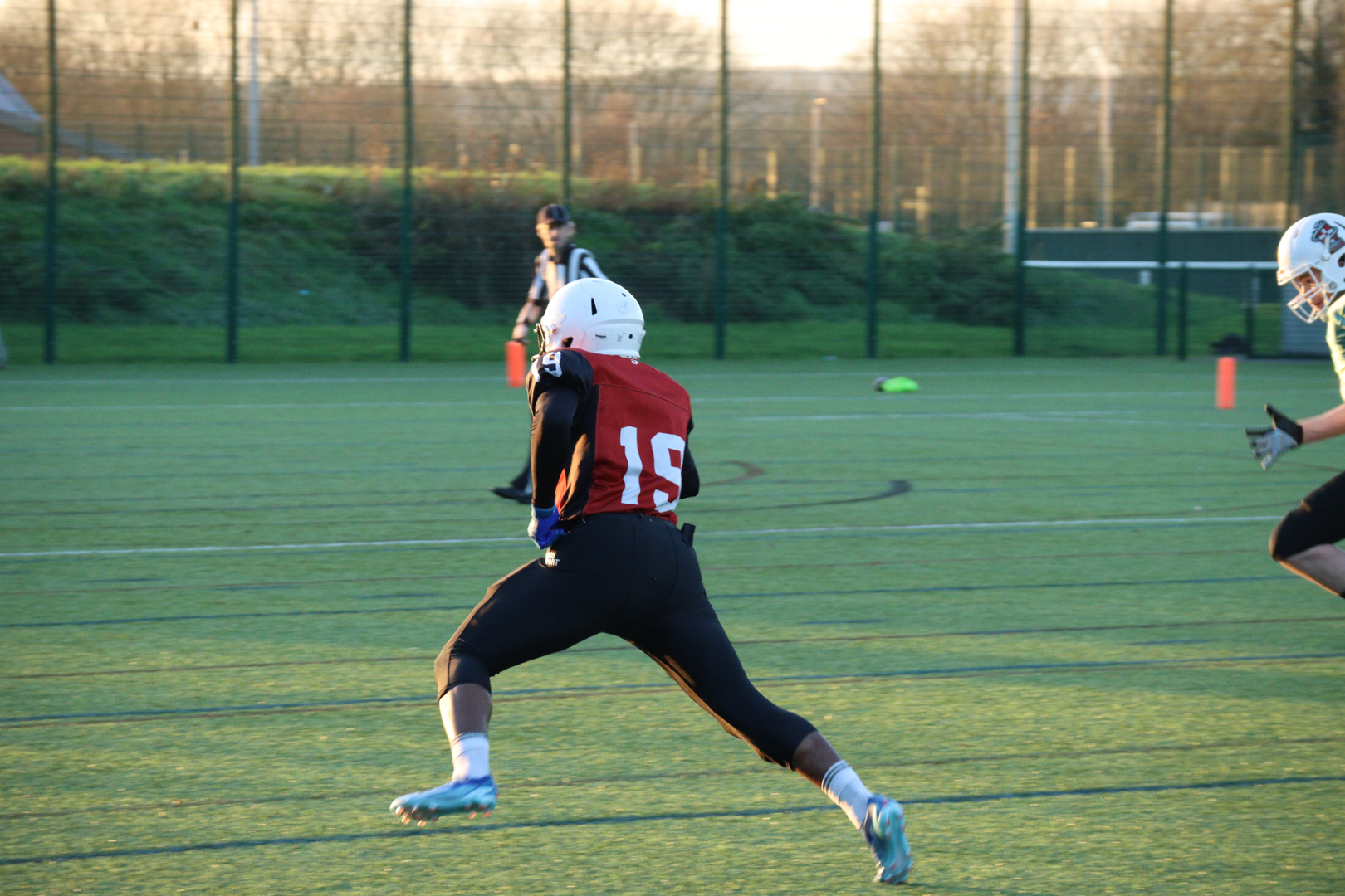 American Football player in a red and black uniform runs on a field, holding a ball, with another player in pursuit.