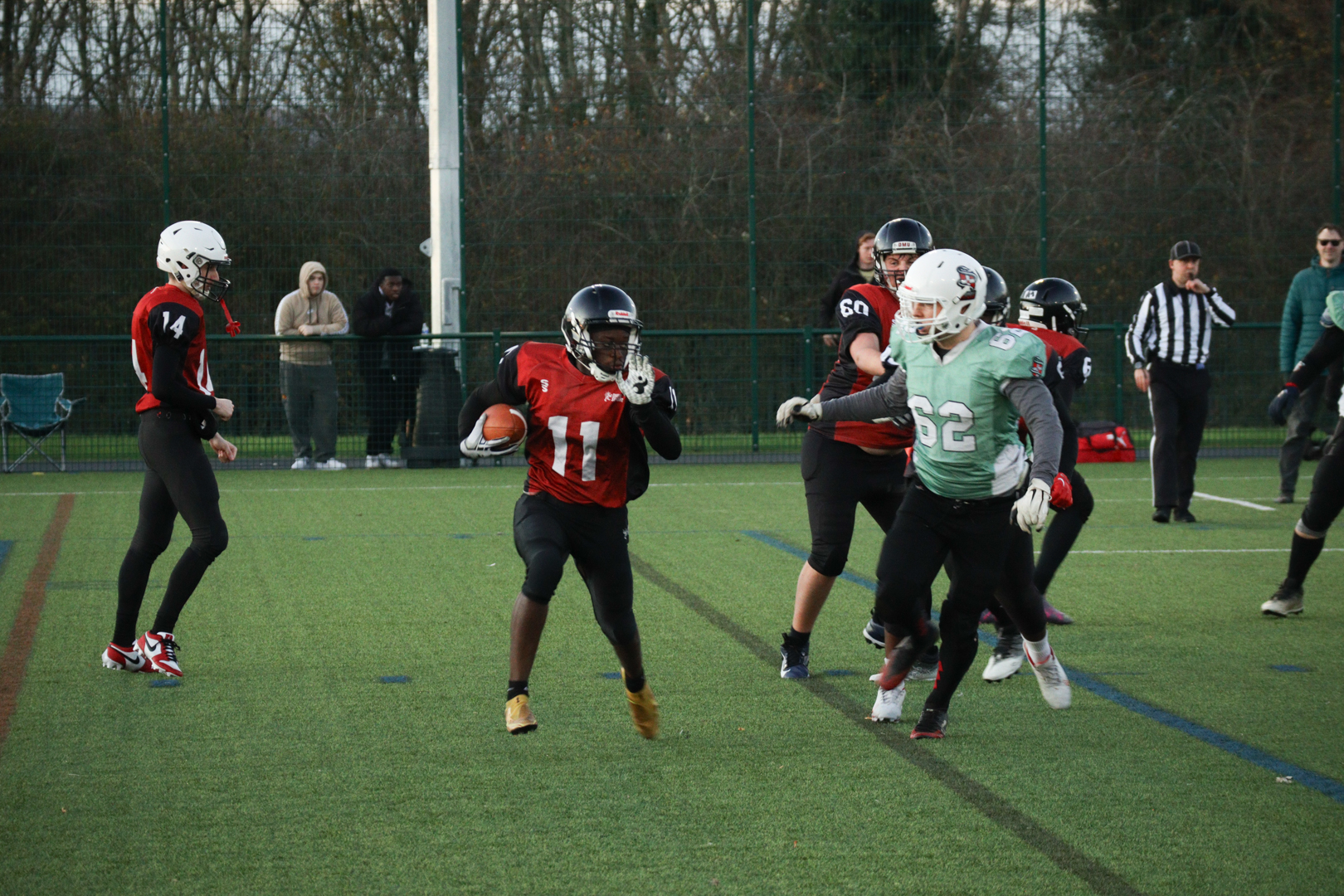 American Football players in red and black uniforms compete, with one player running while holding the ball.