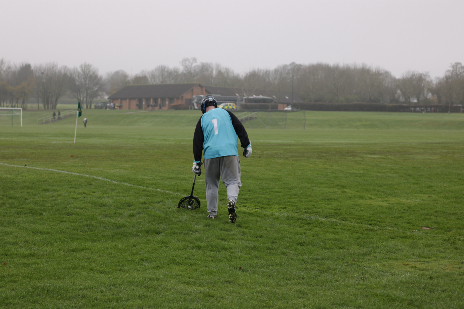 Lacrosse goalkeeper on a grassy field picking up ball.