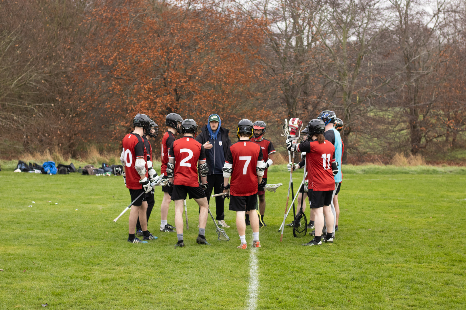 Student lacrosse team in red jerseys huddles with coach on a grassy field, preparing for a game.