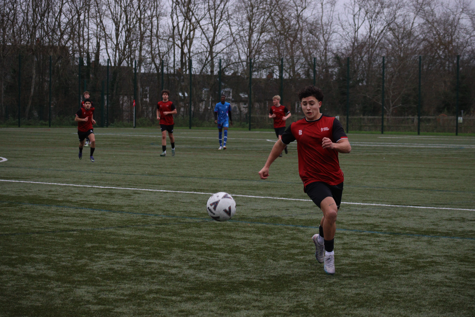 Football player in red shirt running toward a ball on a field with teammates in the background.