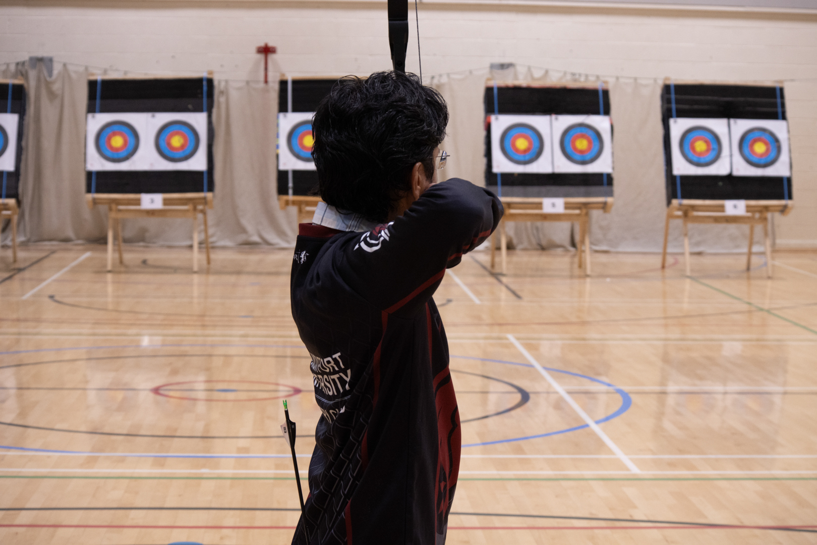Person aiming a bow at archery targets in an indoor gymnasium.