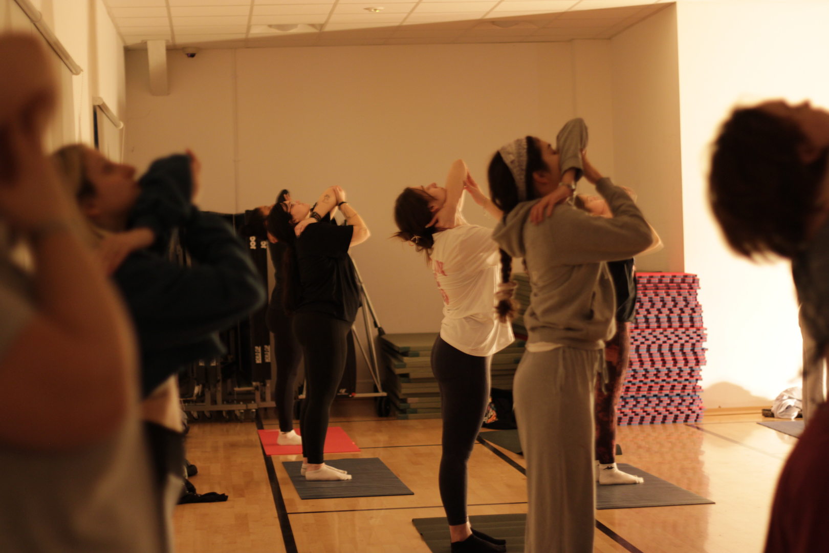 Several people standing on yoga mats in a room, all practicing a pose with arms raised overhead.