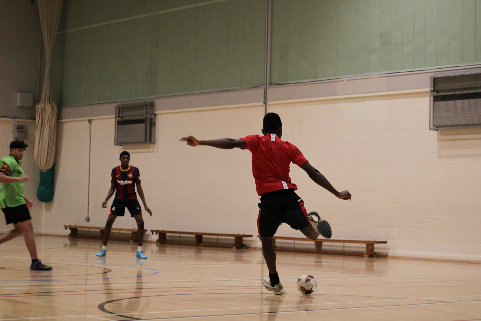 Three people playing indoor football, with one person in a red shirt kicking the ball while others watch or defend.