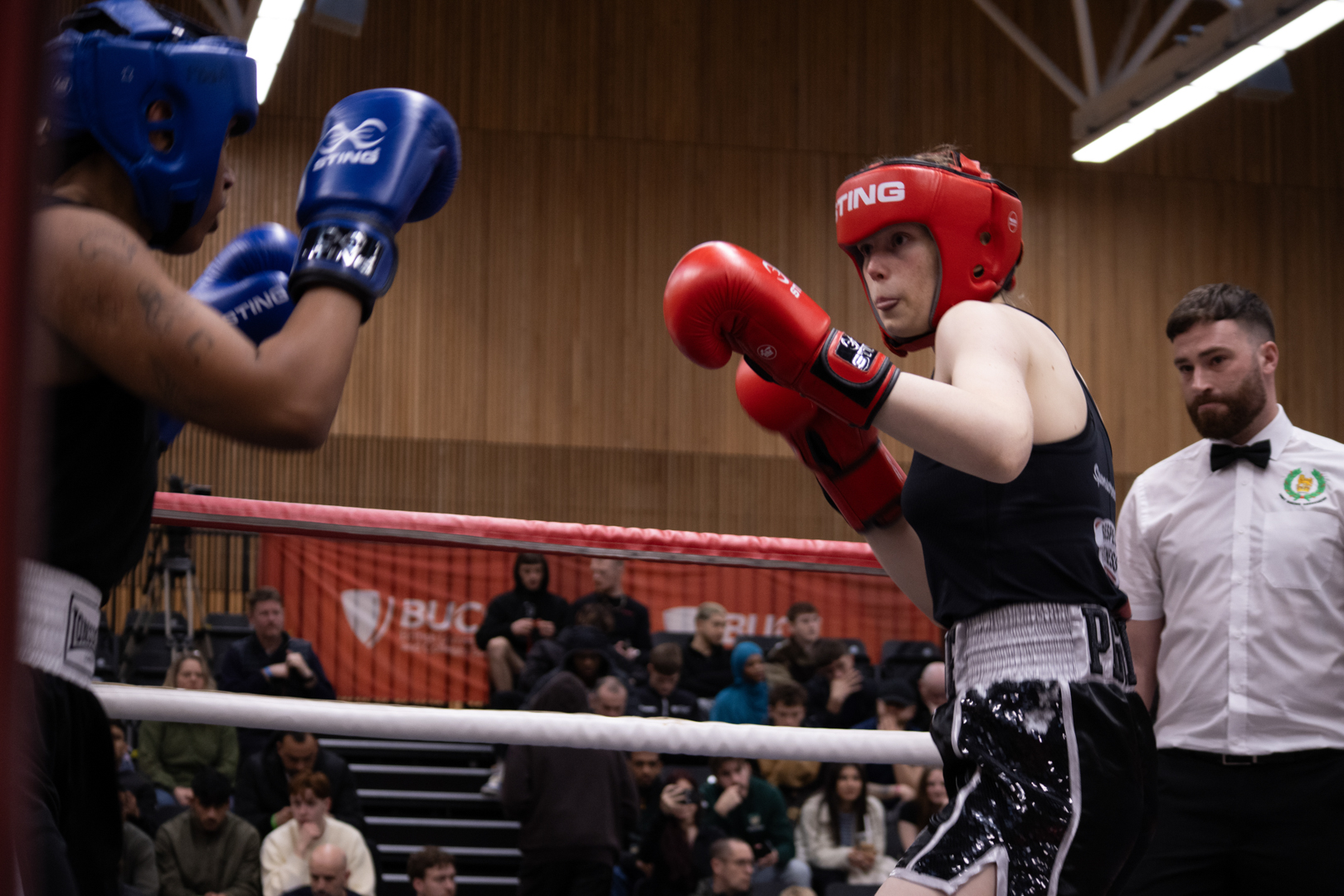 Two boxers in a ring face each other, wearing protective gear and gloves, with a referee observing nearby.